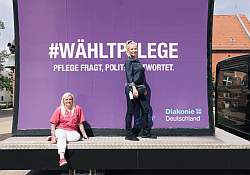 Zwei Pflegekräfte des Ernst-Berendt-Haus der Stephanus-Stiftung stehen und sitzen vor dem Typo-Plakat zur Diakonie-Aktion 'Pflege fragt, Politik antwortet.'